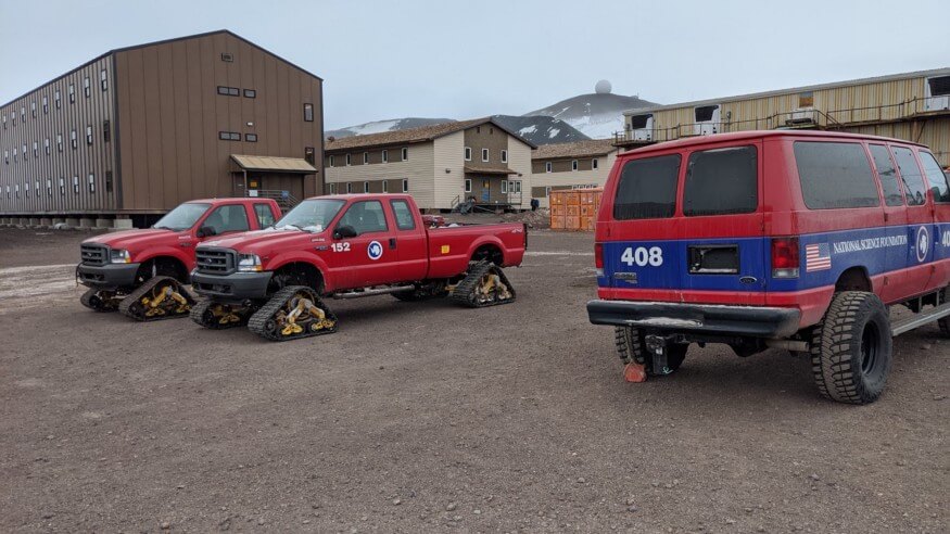 An image of modified track-pickups, and lifted Ford F350 vans in front of McMurdo Station’s Residence Halls.